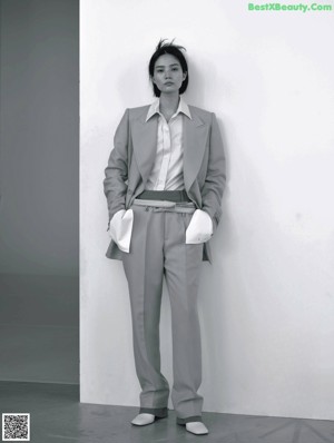 A woman in a beige coat standing in front of a white wall.