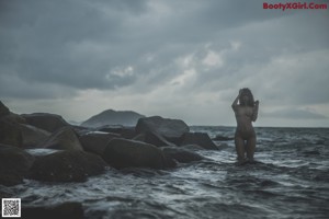 A naked woman standing on a rock by the ocean.